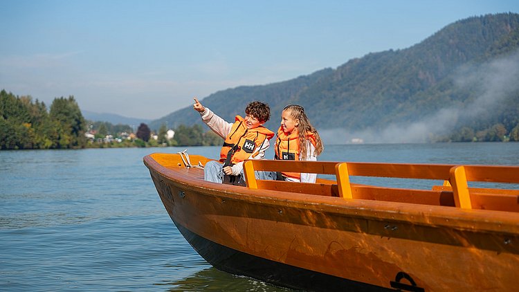 Der Junge zeigt seiner Schwester begeistert etwas, das er während der Zillenfahrt bei der Zillenvermietung Freizell entdeckt hat, während beide Kinder jeweils eine Schwimmweste tragen und über die malerisch blaue Donau gleiten.