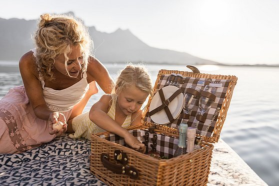 Foto Oberösterreich Tourismus/MondseeIrrsee/Michael Grössinger: Ein Mädchen greift in einen Picknickkorb welcher auf einem Holzsteg am Mondsee im Salzkammergut steht.