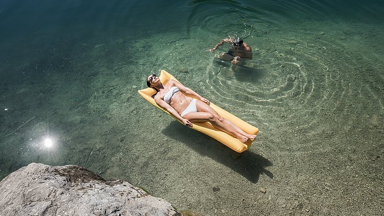 Eine Dame in weißem Bikini liegt auf einer gelben Luftmatratze, die im klaren Wasser schwimmt, und genießt die Sonne. Neben ihr taucht ein Mann im klaren Wasser unter.