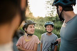 Eine Familie mit zwei Kindern in Nahaufnahme macht gerade eine Pause bei ihrer Radtour am Trattnachtal-Radweg in der Urlaubsregion Vitalwelt Bad Schallerbach. Beide Kinder blicken fröhlich den Vater an, alle tragen einen Radhelm, die Mutter ist im Vordergrund unscharf zu sehen. Im Hintergrund ein Feld im Sonnenlicht.
