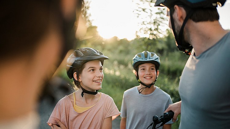 Eine Familie mit zwei Kindern in Nahaufnahme macht gerade eine Pause bei ihrer Radtour am Trattnachtal-Radweg in der Urlaubsregion Vitalwelt Bad Schallerbach. Beide Kinder blicken fröhlich den Vater an, alle tragen einen Radhelm, die Mutter ist im Vordergrund unscharf zu sehen. Im Hintergrund ein Feld im Sonnenlicht.