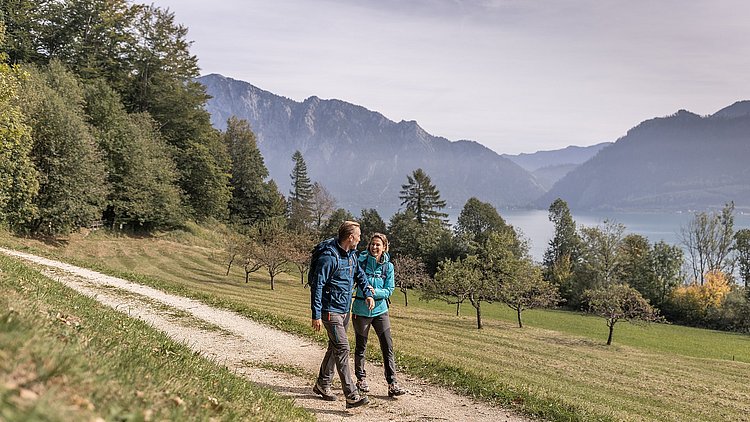 Ein Mann und eine Frau wandern am Edelkastanienweg am Attersee im Salzkammergut, die Frau hält den Mann am Arm, beide lachen, im Hintergrund Berge und der Attersee.