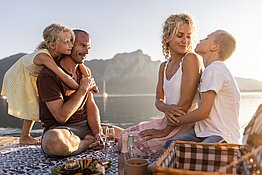 Familie in der Abendsonne beim Picknick auf einem Steg am Mondsee im Salzkammergut, alle sitzen auf einer Picknickdecke, im Hintergrund der Mondsee.