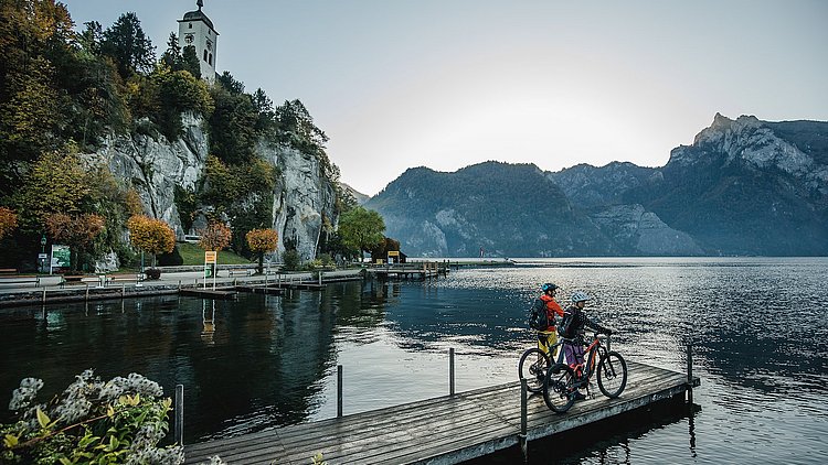Eine Frau und ein Mann stehen am Steg mit dem Fahrrad. Die beiden blicken direkt auf die Kirche in Traunkirchen am Traunsee.