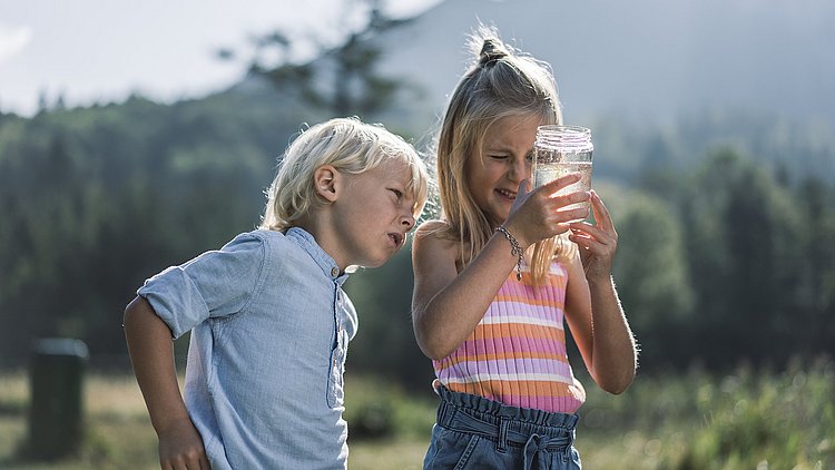 Zwei Kinder in Grünberg im Salzkammergut begutachten ein mit Wasser gefülltes Einmachglas. Im Hintergrund ist undeutlich die gründe Landschaft zu sehen.