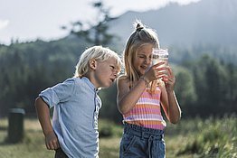 Zwei Kinder in Grünberg im Salzkammergut begutachten ein mit Wasser gefülltes Einmachglas. Im Hintergrund ist undeutlich die gründe Landschaft zu sehen.