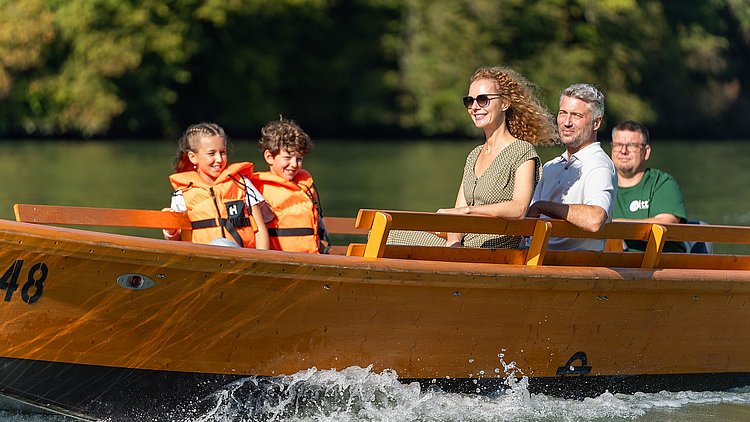 Eine Familie fährt mit einer Holzzille über die Donau, links sitzen der Junge und seine Schwester mit Rettungswesten gesichert, gegenüber das Elternpaar, während im Heck Gerald Witti von der Zillenvermietung Freizell die Zille souverän und ruhig steuert.