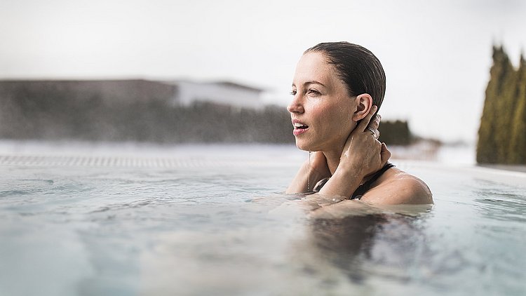 Eine Frau ist im Winter bis zum Hals in das Wasser eines dampfenden Outdoor-Pools im Spa Resort Therme Geinberg im Innviertel eingetaucht.