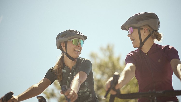 Zwei Frauen, die auf einem Gravelbike sitzen, lachen sich gegenseitig an.