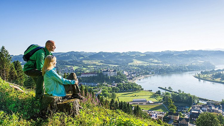 Ein Paar verweilt auf einem schmalen Pfad des Donausteigs und lässt den Blick über die beeindruckende Landschaft schweifen. Unter ihnen glitzert das Wasser der Donau, während die Silhouette von Grein mit dem Schloss Greinburg malerisch hervorsticht.