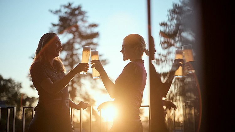 Zwei Frauen stehen auf einer Terrasse. Sie stoßen mit einem Bier an.