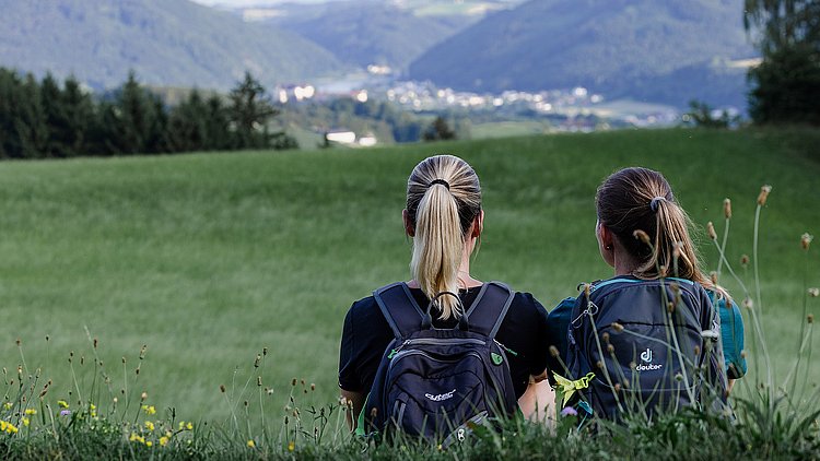 Zwei Freundinnen sitzen in der Wiese bei der Speckalm und genießen den herrlichen Ausblick auf Bad Kreuzen. Umgeben von der malerischen Landschaft und der ruhigen Natur lassen sie den Blick über die idyllische Umgebung schweifen und entspannen sich in dieser friedlichen Atmosphäre.