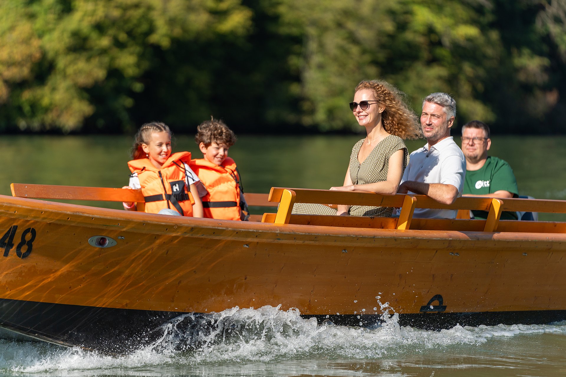 Eine Familie fährt mit einer Holzzille über die Donau, links sitzen der Junge und seine Schwester mit Rettungswesten gesichert, gegenüber das Elternpaar, während im Heck Gerald Witti von der Zillenvermietung Freizell die Zille souverän und ruhig steuert.

