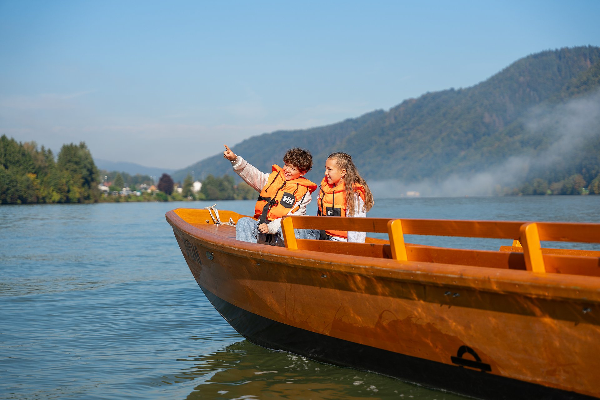 Der Junge zeigt seiner Schwester begeistert etwas, das er während der Zillenfahrt bei der Zillenvermietung Freizell entdeckt hat, während beide Kinder jeweils eine Schwimmweste tragen und über die malerisch blaue Donau gleiten.
