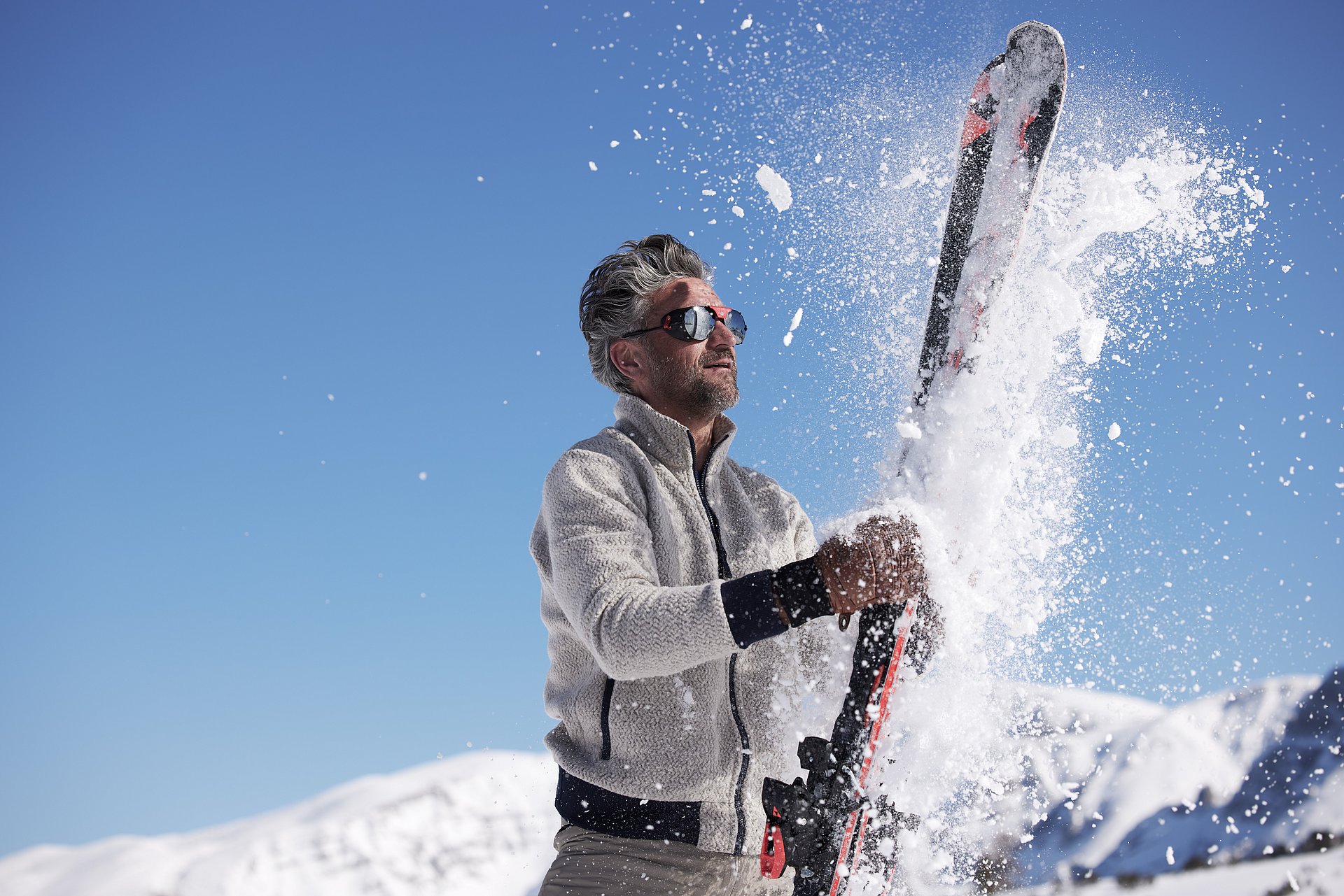 Ein Mann mit weißer Fleecejacke und Sonnenbrille klopft sich im Skigebiet auf der Höss in Hinterstoder den Schnee von seinen Skiern.