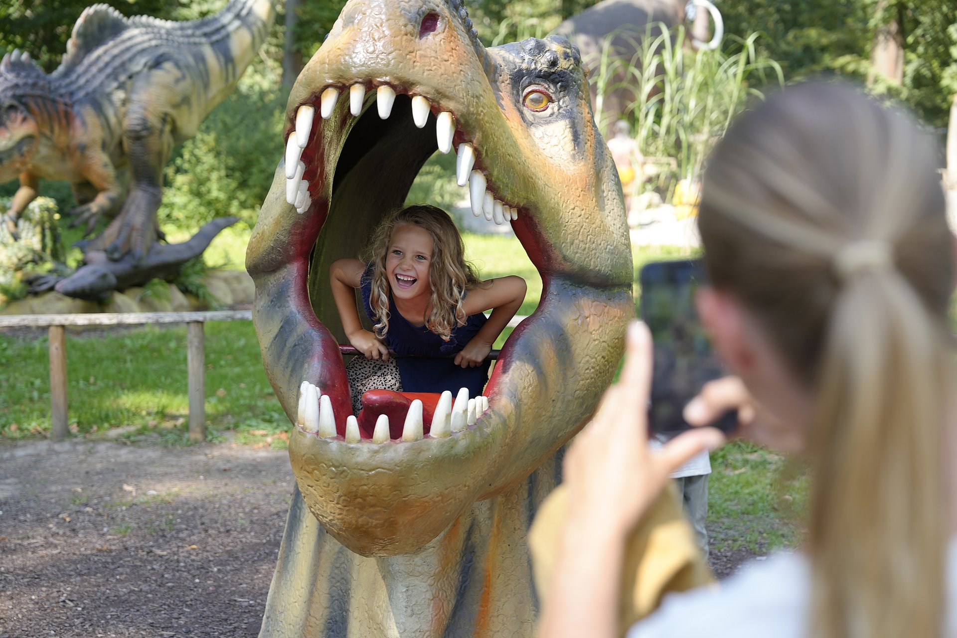 Ein gut gelauntes Mädchen sitzt im geöffneten Maul einer Saurierfigur im Dinoland Schloss Katzenberg im Innviertel.  Im Bildvordergrund ist unscharf eine Frau zu erkennen welche mit ihrem Smartphone ein Bild vom Kind macht.