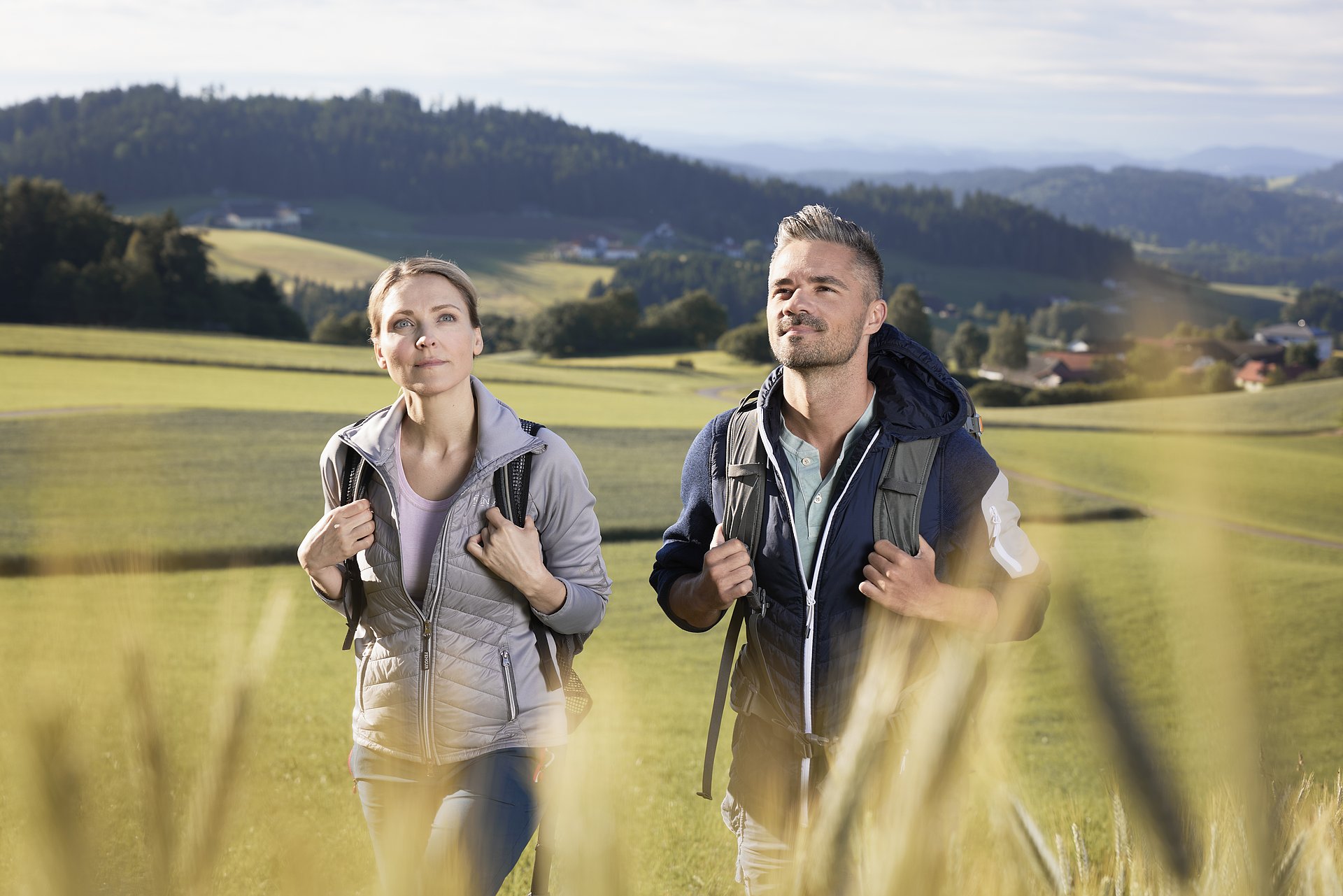 Eine Frau und ein Mann wandern auf einer Wiese in der Region Mühlviertler Alm Freistadt, im Bildhintergrund sind Häuser und die hügelige Landschaft des Mühlviertels zu sehen.
