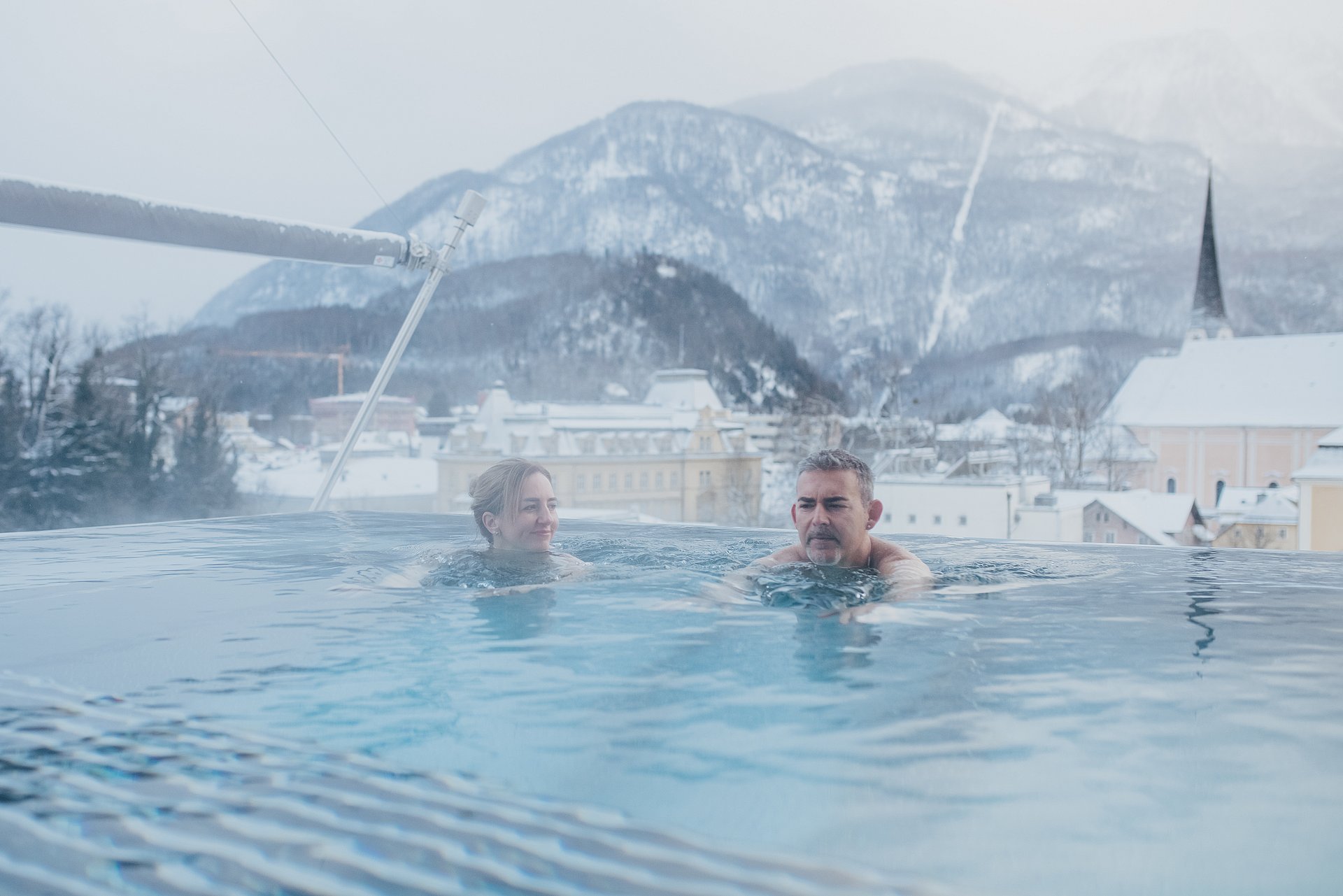 Eine Frau und ein Mann sind in einem Außenpool auf einem Dach im Winter und genießen eine schöne Aussicht auf die Dächer in Bad Ischl.  Es schneit sehr leicht.