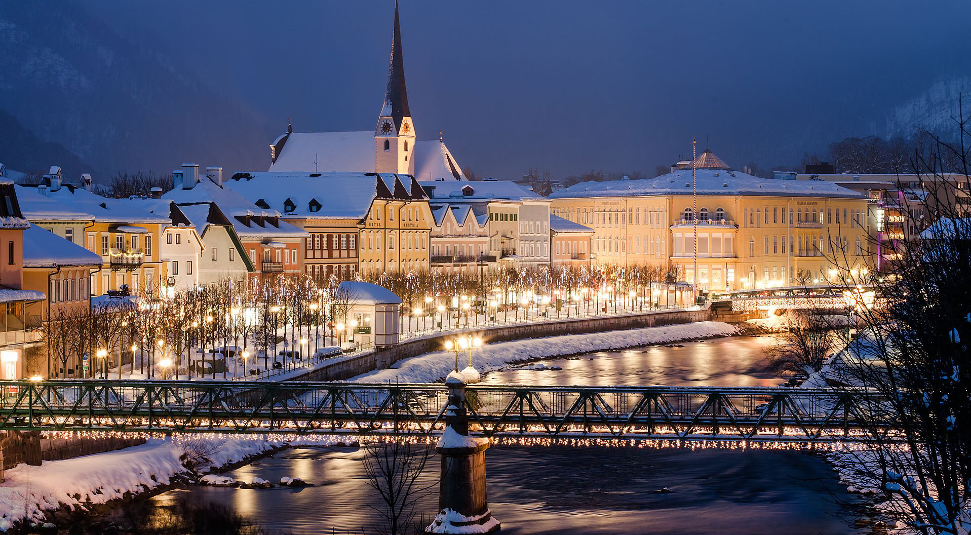 Schneebedecktes Flussufer am Abend, Bruecke im Vordergrund und die Baumallee im Hintergrund sind beleuchtet